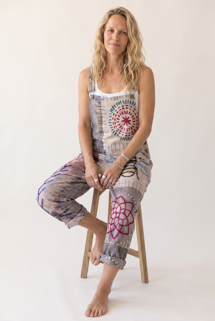 A woman sits on a wooden stool wearing a sleeveless jumpsuit with a white underlayer. The jumpsuit features a multicolor abstract print with circular mandala-like patterns in red, blue, and pink on a beige and gray base.